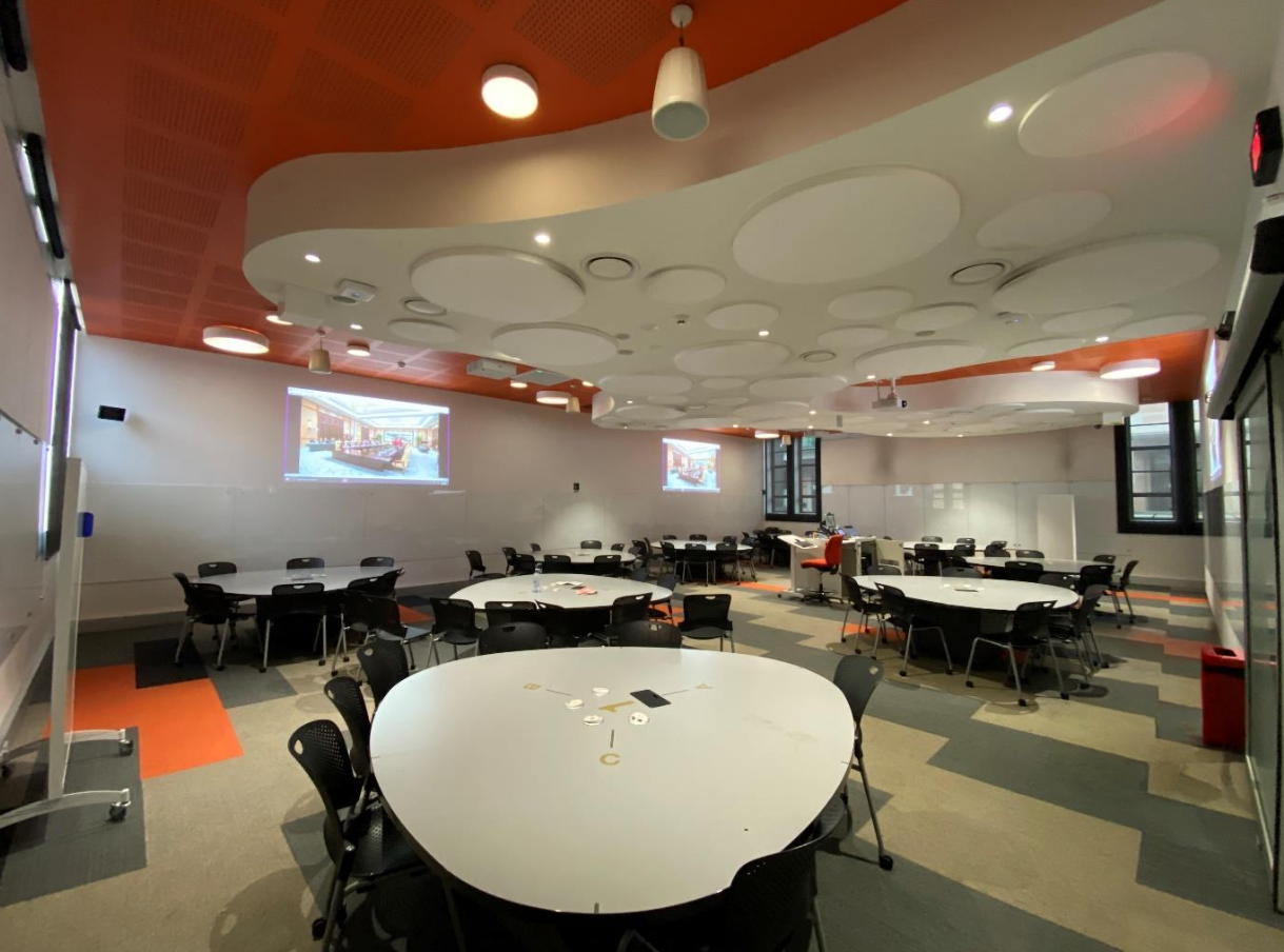 Innomate x University of Queensland: A modern teaching room with advanced technology and ample seating. Projectors, screens, and vibrant orange accents on the ceiling highlight the innovative learning environment.